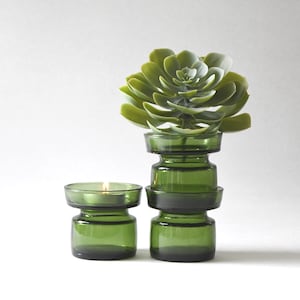 Può includere: Due portacandele in vetro verde, uno con una candela accesa, e un set impilato di tre portacandele in vetro verde sormontati da una pianta succulenta verde. I portacandele hanno un design semplice e moderno.