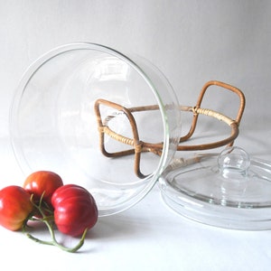 May include: A clear glass bowl with a matching glass lid, resting on a light brown woven rattan stand with two handles. Three red tomatoes with green stems are positioned in front of the glass bowl on a plain white surface.