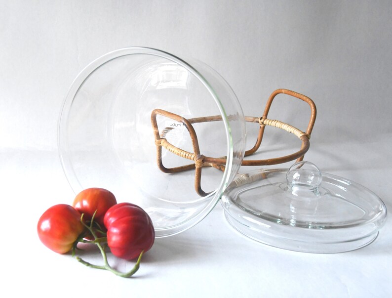 May include: A clear glass bowl with a matching glass lid, resting on a light brown woven rattan stand with two handles. Three red tomatoes with green stems are positioned in front of the glass bowl on a plain white surface.