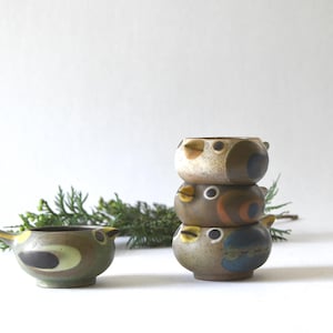Può includere: Quattro ciotole in ceramica a forma di uccello, caratterizzate da smalti maculati marroni, beige e blu con occhi e becchi dipinti. Una ciotola è posizionata da sola a sinistra, mentre tre sono impilate verticalmente a destra, con corpi arrotondati.
