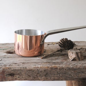 May include: A copper saucepan with a long, silver handle. The interior is silver, and the exterior is a shiny copper. The pan sits on a weathered wooden surface, with a pinecone and twigs nearby. Kitchenware.