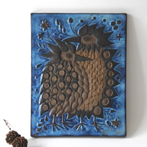 Puede incluir: Azulejo de pared de cerámica con un diseño estilizado de dos pájaros marrones y negros sobre fondo azul. El azulejo es de forma rectangular y está decorado con elementos florales. Medidas en centímetros.