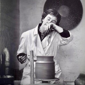 May include: A woman in a white coat is working on a pottery wheel. She is holding a piece of clay in her hands and is concentrating on her work. The pottery wheel is made of wood and has a metal base. The woman is wearing a necklace and has her hair pulled back.