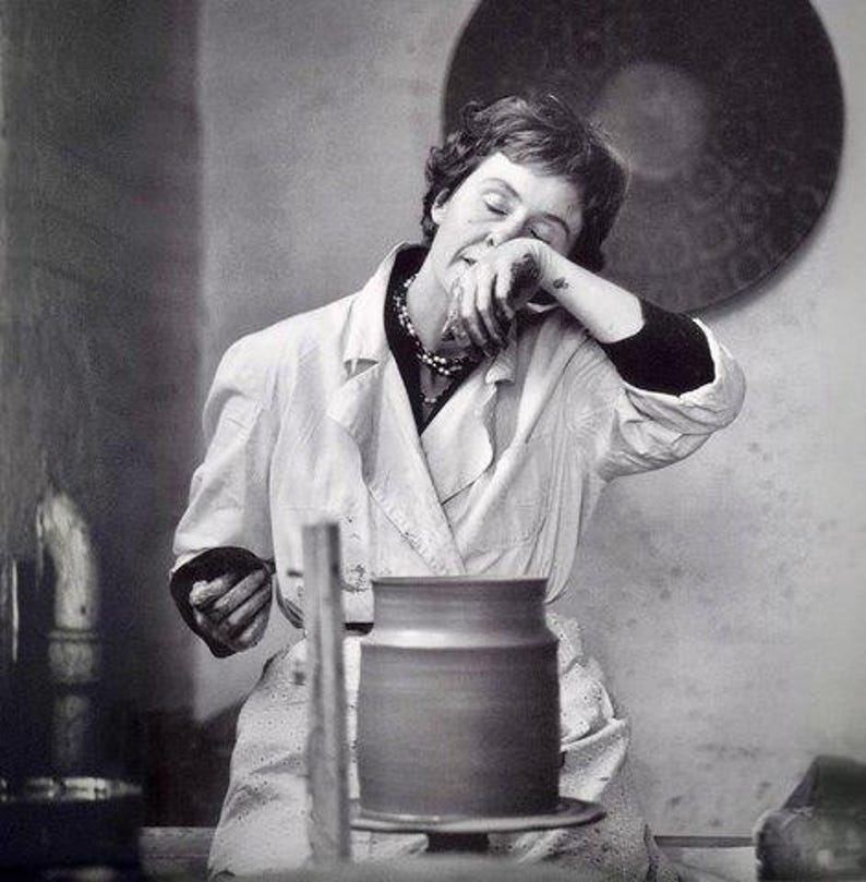 May include: A woman in a white coat is working on a pottery wheel. She is holding a piece of clay in her hands and is concentrating on her work. The pottery wheel is made of wood and has a metal base. The woman is wearing a necklace and has her hair pulled back.