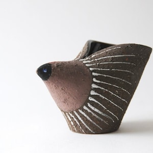 May include: A small, hand-formed ceramic vessel shaped like a stylized bird. The body is dark brown with a textured surface, featuring white vertical etched lines along the sides and a smooth, lighter brown head with a dark, glossy beak tip.
