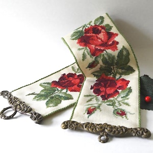 May include: Cross-stitched floral wall hanging featuring red roses and green leaves on an off-white background. The fabric is framed by ornate, antique-style metal hardware. A decorative piece for home decor.
