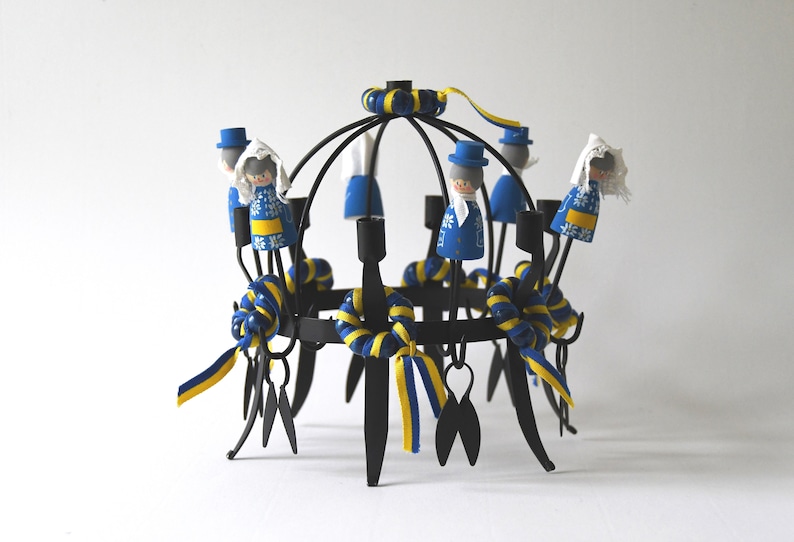 Puede incluir: Un candelabro de metal negro con un dise&ntilde;o circular, adornado con figuras de madera vestidas con trajes tradicionales azules y amarillos. El candelabro presenta cintas decorativas azules y amarillas, sobre fondo blanco.