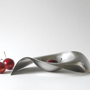 May include: A modern, brushed metal bowl with a flowing, wave-like design. Three red cherries with green stems are placed near the bowl. The bowl is empty except for one cherry.