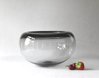 May include: A large, clear glass bowl with a dark gray rim. The bowl is sitting on a white surface with two red strawberries in front of it.