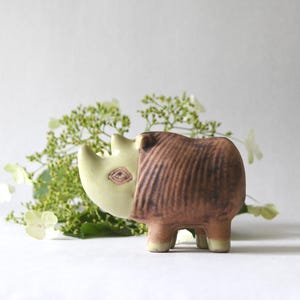 Może przedstawiać: Ceramiczna figurka nosorożca z gładką głową w kolorze jasnego kremu oraz teksturowanym, ciemnobrązowym korpusem z pionowymi żłobieniami. Zwierzę posiada wyraźny róg oraz mały, szczegółowy znak oka po bocznej stronie głowy.
