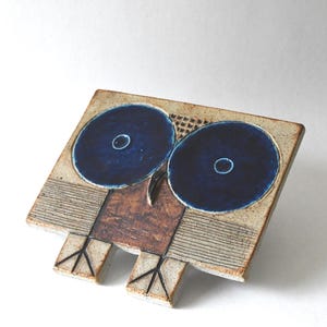 Puede incluir: Escultura de cerámica de un búho con grandes ojos azul oscuro. El búho tiene un cuerpo cuadrado con una superficie texturizada de color beige y detalles marrones. Los ojos son circulares con un centro azul más claro. Las patas del búho están representadas con líneas negras.