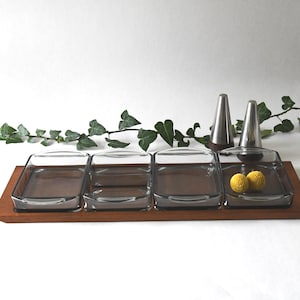 Puede incluir: Una bandeja de servir de madera con cuatro platos de vidrio y dos saleros y pimenteros de plata. La bandeja es marrón y los platos son transparentes. Hay dos botones de flores amarillas en uno de los platos.