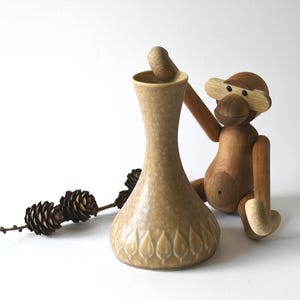 Può includere: Un vaso in ceramica beige con un motivo a foglie, una statuina di scimmia in legno e un rametto di pigne. Il vaso è alto circa 20 cm. La scimmia ha arti mobili. Gli oggetti sono su sfondo bianco.