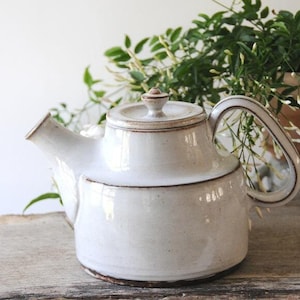 Puede incluir: Una tetera de cerámica blanca con un borde marrón y un asa marrón. La tetera tiene un cuerpo redondeado y una tapa con un pomo en la parte superior.