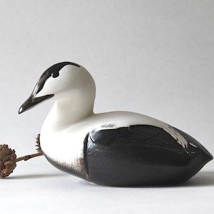 May include: A ceramic duck figurine with black and white markings. The duck is sitting on a white surface with a small brown pinecone next to it.