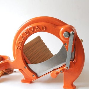 Puede incluir: Cortadora de alimentos de hierro fundido naranja con una tabla de cortar de madera y una cuchilla plateada. La cortadora está etiquetada como "BAADVAAD".