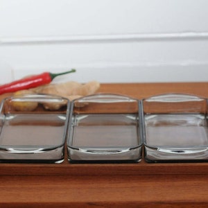 Puede incluir: Tres fuentes rectangulares de vidrio transparente con asas curvas sobre una bandeja de madera. Un pimiento rojo y jengibre están sobre la superficie de madera. Las fuentes están vacías y listas para usar.