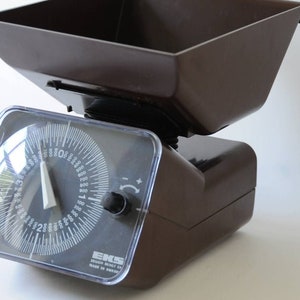 Op de afbeelding: Een bruine vintage keukenweegschaal met een heldere plastic voorkant en een ronde wijzerplaat. De weegschaal heeft een rechthoekige basis en een bruine kom bovenop. De wijzerplaat toont metingen tot 1000 gram.