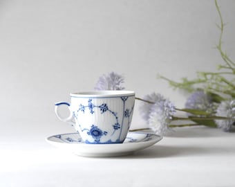 Puede incluir: Un juego de taza de té y platillo de porcelana blanca con un delicado patrón floral azul. La taza presenta una textura acanalada y una pequeña asa, mientras que ambas piezas están decoradas con intrincadas enredaderas y flores azules.