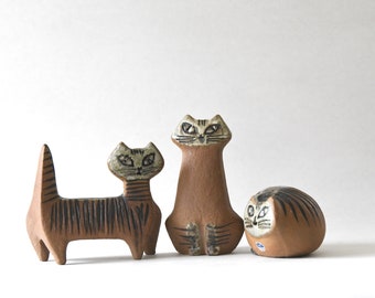 May include: Three terracotta cat figurines with hand-painted black striped patterns. One cat stands with a raised tail, one sits upright, and one is curled in a rounded shape. Each figure features a light-colored face with detailed black eyes.