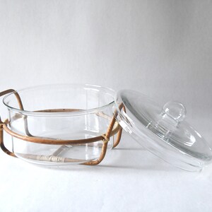 May include: A clear glass casserole dish with a matching glass lid featuring a round knob handle. The dish is held within a light brown woven rattan frame that includes two side handles and a base support structure for the circular glass vessel.