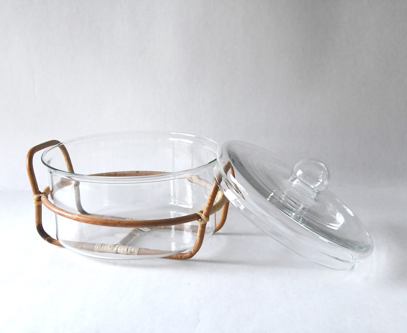 May include: A clear glass casserole dish with a matching glass lid featuring a round knob handle. The dish is held within a light brown woven rattan frame that includes two side handles and a base support structure for the circular glass vessel.