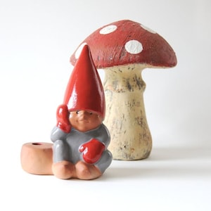 May include: A ceramic gnome figurine wearing a tall red hat and grey clothing with red mittens, positioned next to a terracotta-colored candle holder and a large decorative mushroom with a red cap featuring white spots and a textured beige stem.