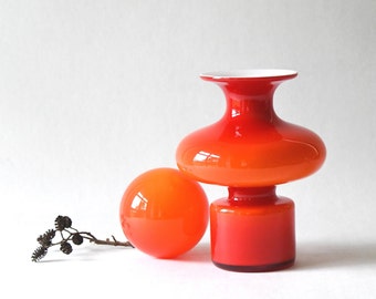Op de afbeelding: Een rode en oranje glazen vaas met een witte rand en een ronde, oranje glazen bol. De vaas heeft een brede, ronde basis en een smalle hals.