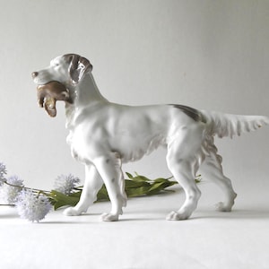 May include: A white porcelain dog figurine with brown markings, holding a brown object in its mouth. The dog is standing next to a sprig of green leaves and several light purple flowers. The figurine has a glossy finish.