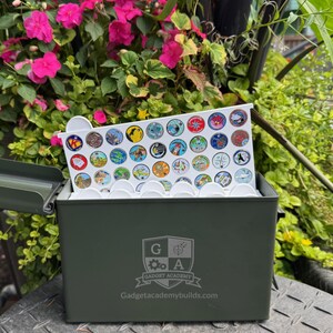 May include: An olive green metal storage box with a white tray filled with colorful round tokens. The tokens have various designs. The box has the text "Gadget Academy" and "Gadgetacademybuilds.com" printed on the front.