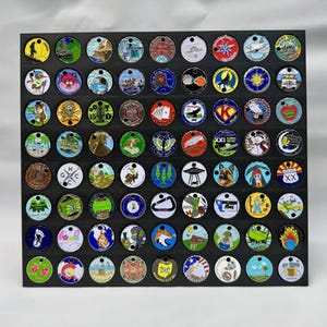May include: A collection of colorful round metal coins with various designs, displayed on a black grid. The coins feature images of landscapes, animals, and symbols. The grid is made of black wood.