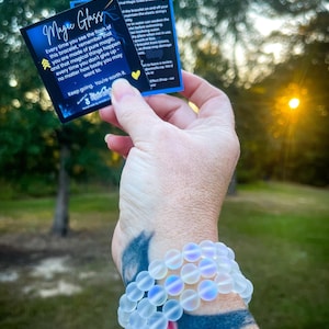 May include: A white glass bead bracelet with iridescent accents. The bracelet is on a person's wrist. The image also includes two cards with text about the bracelet. The text on the cards reads "Magic Glass" and "Viral Magic Class Bracelet".