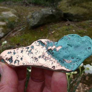 May include: A handcrafted copper sculpture with a cloud-like shape. The sculpture is partially oxidized, displaying a turquoise patina on one side and a polished copper finish on the other. The piece is held in a hand.