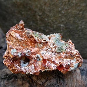May include: A raw copper specimen with a rough, textured surface. The copper displays a mix of reddish-brown and orange hues, with patches of green and white mineral deposits. The natural formation is displayed on a piece of wood.
