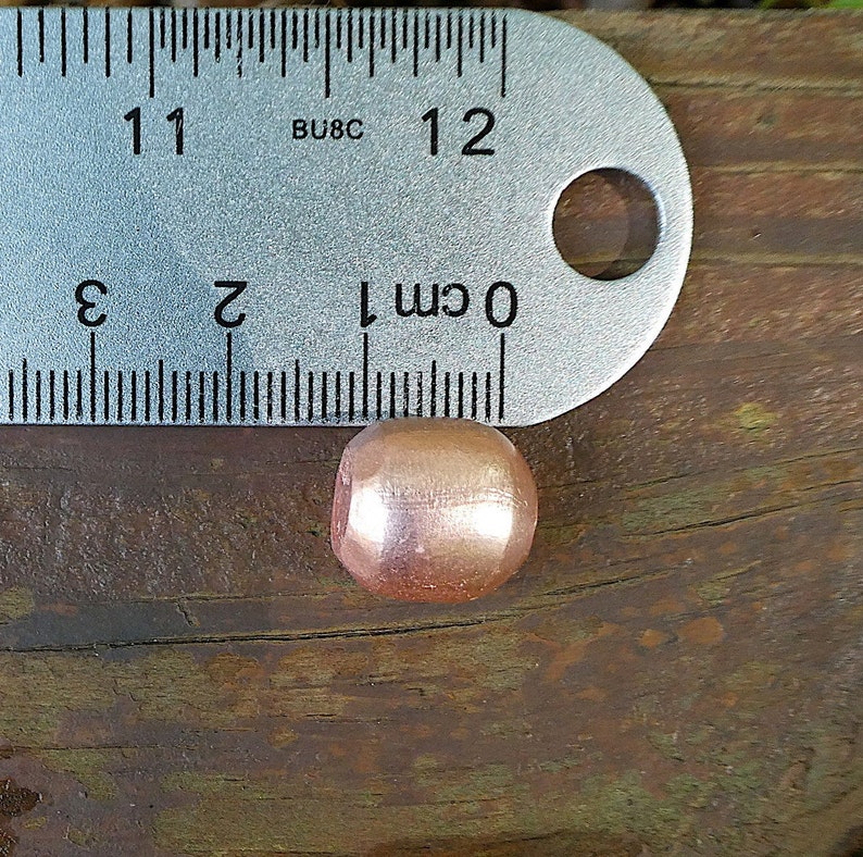 K&ouml;nnte beinhalten: Nahaufnahme einer einzelnen, runden, metallischen Perle mit gl&auml;nzender ros&eacute;goldener Oberfl&auml;che. Die Perle hat einen Durchmesser von etwa 1 cm und liegt auf einer Holzoberfl&auml;che.