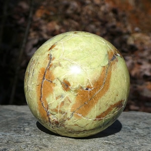 May include: A spherical, polished gemstone with a light green hue, veined with brown and dark lines. The orb is smooth and reflective, showcasing natural patterns. The stone is likely a decorative item or a collectible.