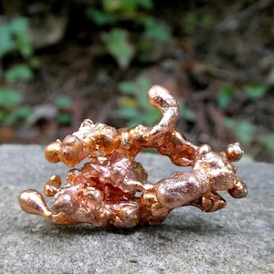 May include: A raw, metallic copper nugget with an irregular, organic shape featuring multiple rounded protrusions and a shiny, reddish-brown surface texture. The piece displays a complex, porous structure with various bumps and curves.