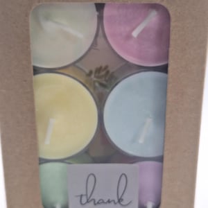 Könnte beinhalten: Eine Schachtel mit sechs pastellfarbenen Teelichtern. Die Kerzen sind in einem Kreis angeordnet und von einer braunen Pappbox umgeben. Die Schachtel hat ein kleines Fenster, das die Kerzen zeigt. Die Schachtel hat einen Aufkleber mit der Aufschrift "Danke".
