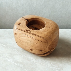 May include: A natural wood candle holder with a rounded, organic shape. The light brown wood grain is visible, with a central circular opening for a candle. The holder has several small, drilled holes and a crack on the side.