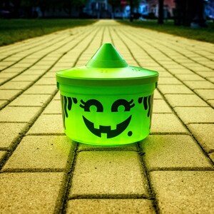 May include: A bright green Halloween candy bucket with a conical lid and a smiling jack-o'-lantern face. The bucket is sitting on a brick walkway, perfect for trick-or-treating or Halloween decorations.