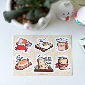 May include: A sheet of six illustrated stickers featuring cute cartoon bread characters. The stickers feature phrases like "Carb Crab", "Sunny Side Up", "Bready or Not", "All You Knead is Love", "Live Laugh Loaf", and "Toastea".