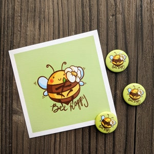 Könnte beinhalten: Eine grüne Karte mit einer Cartoon-Biene, die eine Blume hält, und dem Text "Bee Happy". Es gibt drei runde Knöpfe mit dem gleichen Bienendesign und dem Text "Bee Happy" auf jedem.