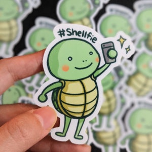 Könnte beinhalten: Eine grüne Schildkröte mit einem gelben Panzer hält ein schwarz-weißes Telefon und lächelt. Die Schildkröte macht ein Selfie und der Text #Shellfie befindet sich über der Schildkröte.