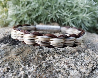 May include: Braided bracelet made of horse hair in shades of brown and white. The bracelet is secured with a silver-colored magnetic clasp. The braided design is visible, showcasing the different colors of the hair.
