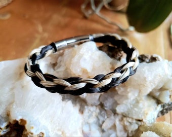 May include: Braided bracelet featuring black and light tan strands. The bracelet has a magnetic clasp and is displayed on a white, crystalline surface. The bracelet's woven design is visible, showcasing the contrast between the dark and light strands.