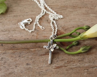 May include: A silver chain necklace with a silver cross pendant featuring a butterfly design. The necklace is lying on a wooden surface with a green plant.