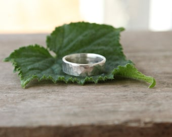 May include: A silver hammered band ring resting on a green leaf.