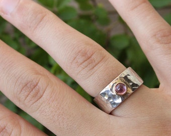 May include: A silver ring with a hammered texture and a pink gemstone set in the center. The ring is worn on a finger.