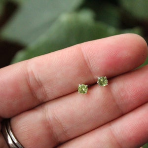 May include: A pair of small, round stud earrings with a light green gemstone. The earrings are set in a silver metal. The earrings are displayed on a hand, with a blurred green background.