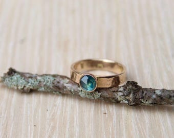 May include: A gold ring with a round, teal-colored gemstone. The ring is set on a textured band. The ring is displayed on a small, lichen-covered branch against a light wood background. The ring is a piece of jewelry.